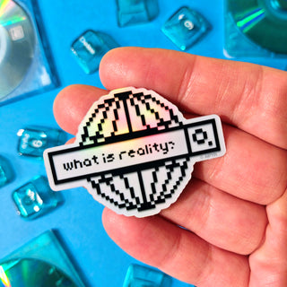 Hand holding a pixelated sticker with 'what is reality?' text against a blue background with transparent crystals.