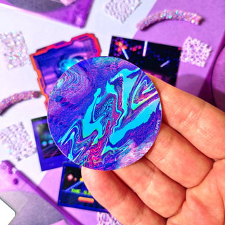 Holographic sticker held by a hand with a colorful, abstract design on a purple background.