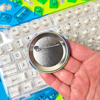Pinback button displayed on a human hand to show the pin mechanism on the back - held over a clear keyboard on a blue and green background.