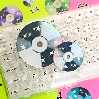 Two star-patterned CDs on a transparent keyboard with colorful CD cases in the background.